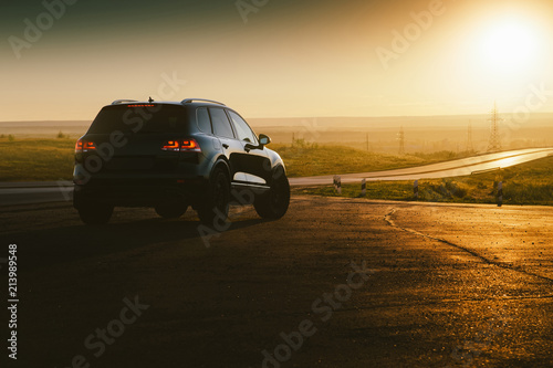 Black car is parked at countryside asphalt road near highway at golden sunset