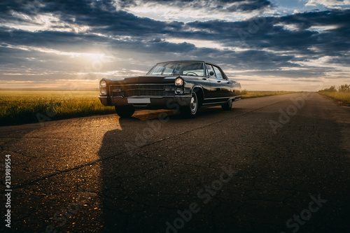 Black retro vintage muscle car is parked at countryside asphalt road at golden sunset © Ivan Kurmyshov