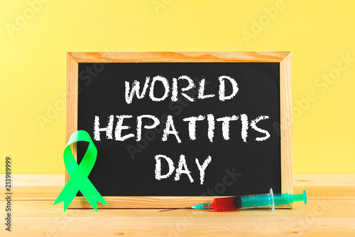Fototapeta Naklejka Na Ścianę i Meble -  Chalkboard with text World Hepatitis Day. June 28th. A green jade ribbon and a syringe with blood on a wooden table against the background of a yellow wall.