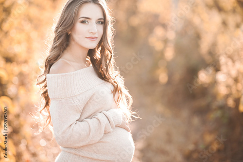 Smiling pregnant woman wearing cozy knitted sweater over autumn background. Looking at camera. Motherhood. Maternity. 20s.