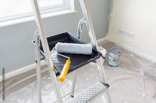 Painting and decorating with a roller and tray on a set of metal step ladders with cans of paint and a dust sheet in the background photo