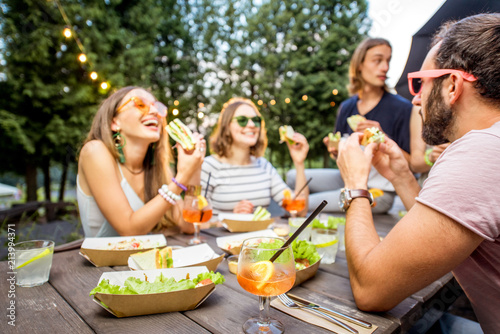 Young friends having fun sitting together with tasty snacks and drinks during the evening lights at the park cafe outdoors photo
