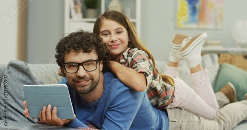 Portrait of the handsome father in glasses with tablet device lying on the sofa and his little cute daughter lying on his back, then they smiling to the camera. Inside. photo