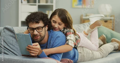 Portrait of the cheerful caucasian father lying on the sofa at home, talking with his little daughter on his back while they watching something on the tablet computer. Indoors. photo