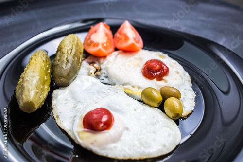 perverted breakfast, fried eggs in the form of nipples, olives, canned cucumbers, tomatoes and ketchup. black ceramic dishes on a matte board.