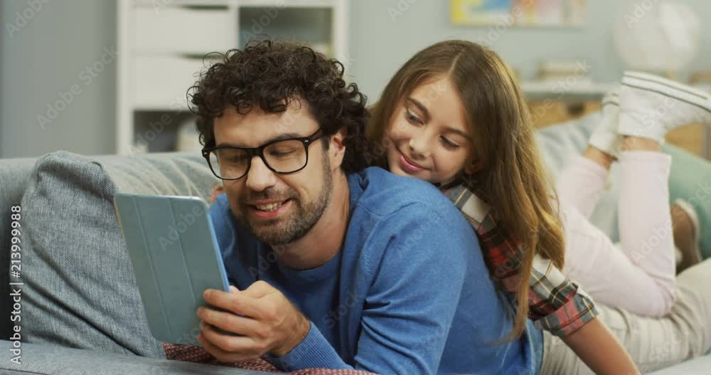 Close up of the joyful smiled man lying together with small daughter on the couch in the living room while they watching something on the tablet device or playing a game. Inside. Close up of the joyful smiled man lying together with small daughter on the couch in the living room while they watching something on the tablet device or playing a game. Inside.