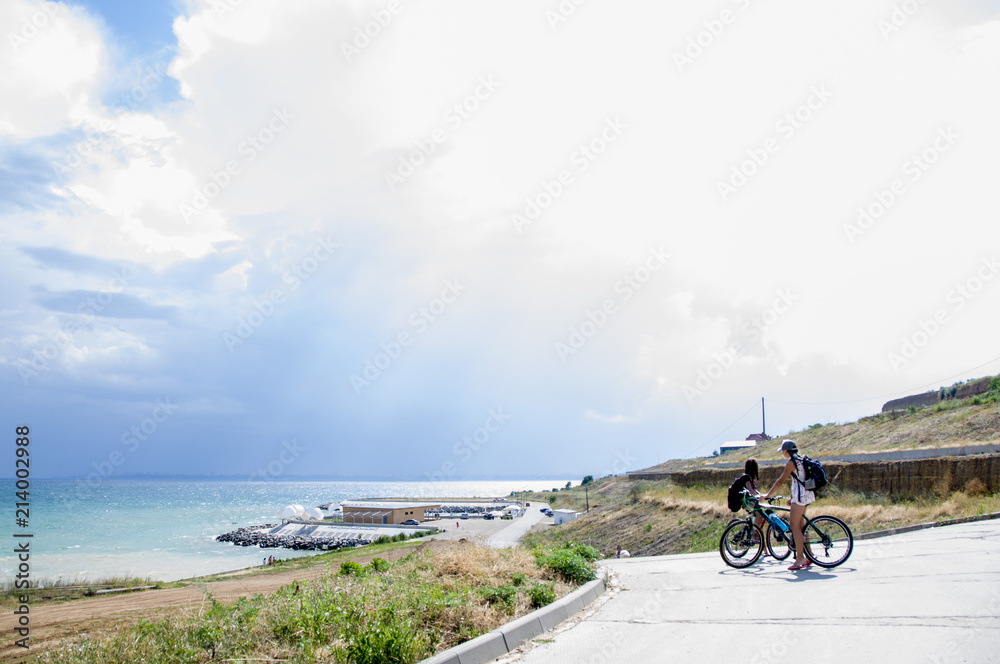 two teenage girls on bicycles on a concrete path to the sea in summer. Water landscape with clouds on the horizon. Sea nature of tranquility. The weather is changing.