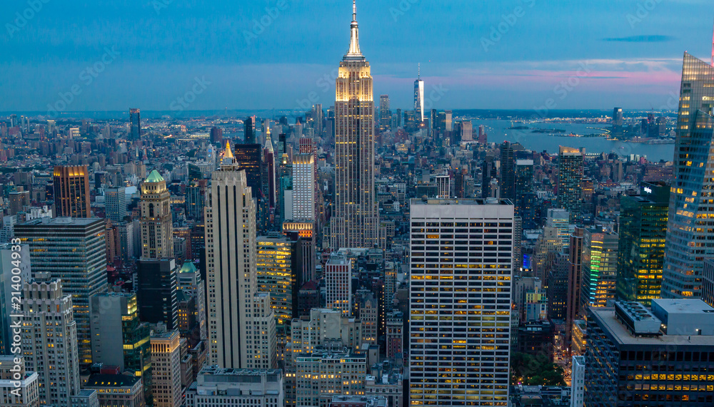 View of Manhattan skyline during the blue hour or twilight. Golden lights in buildings, surrounded by brilliant blue sky with touches of pink on horizon.