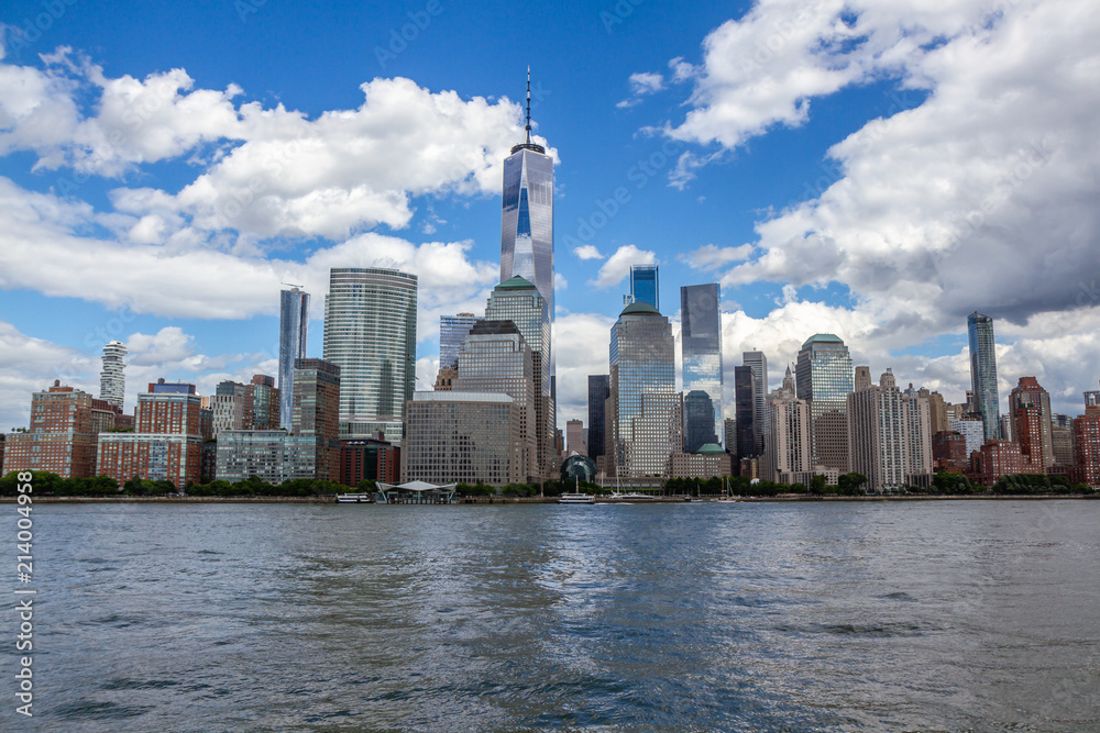 View of downtown Manhattan from the water. Freedom Tower in middle of frame, white clouds in blue sky.