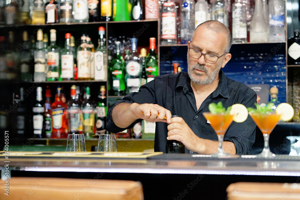 A bartender men smiles and opens a bottle of white wine for the client of the hotel bar. Shelves with bottles of alcohol in the background. The concept of service.
