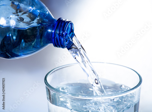 Pouring of cold clear drink water from bottle into glass, close up, blue toned on white background
