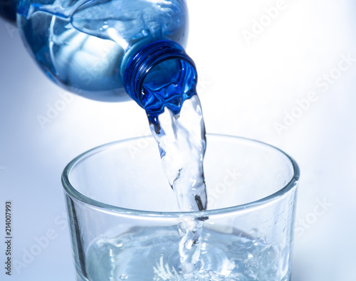 Pouring of cold clear drink water from bottle into glass, close up, blue toned on white background