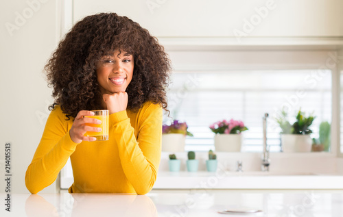 African american woman drinking orange juice in a glass with a happy face standing and smiling with a confident smile showing teeth