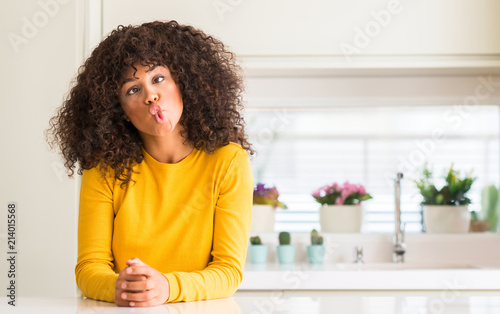 African american woman wearing yellow sweater at kitchen making fish face with lips, crazy and comical gesture. Funny expression.