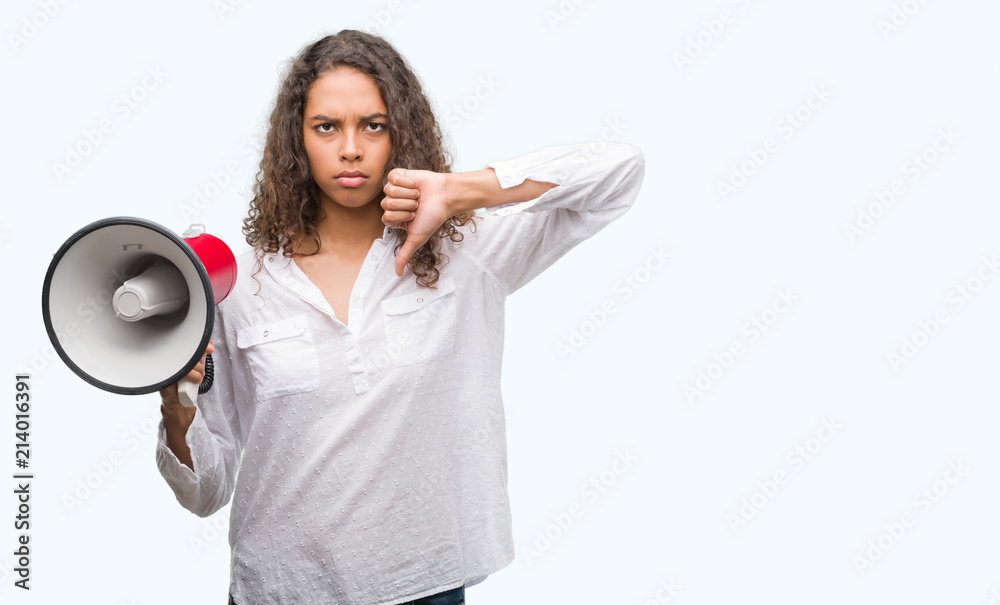 Young hispanic woman holding megaphone with angry face, negative sign showing dislike with thumbs down, rejection concept Young hispanic woman holding megaphone with angry face, negative sign showing dislike with thumbs down, rejection concept