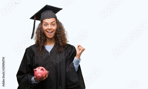 Young hispanic woman wearing graduation uniform holding piggy bank screaming proud and celebrating victory and success very excited, cheering emotion