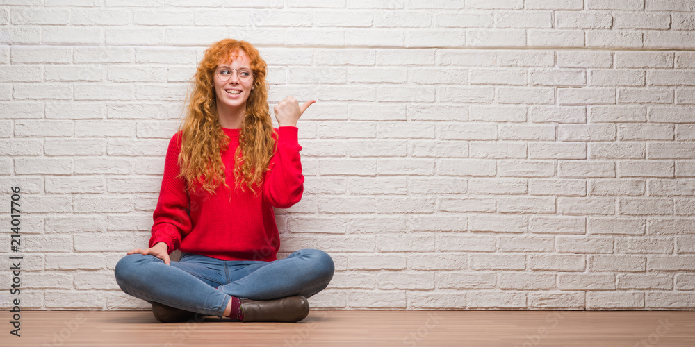 Young redhead woman sitting over brick wall smiling with happy face looking and pointing to the side with thumb up. Young redhead woman sitting over brick wall smiling with happy face looking and pointing to the side with thumb up.