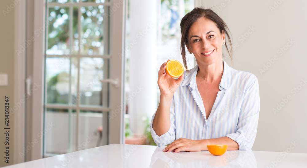 Middle aged woman holding orange fruit with a happy face standing and smiling with a confident smile showing teeth Middle aged woman holding orange fruit with a happy face standing and smiling with a confident smile showing teeth