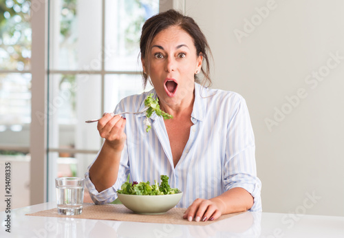 Middle aged woman eating fresh salad in a bowl at home scared in shock with a surprise face, afraid and excited with fear expression