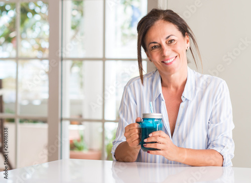 Middle aged woman drinking milk shake in a glass with a happy face standing and smiling with a confident smile showing teeth