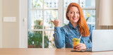 Redhead woman using computer laptop eating fruit at home pointing with hand and finger up with happy face smiling