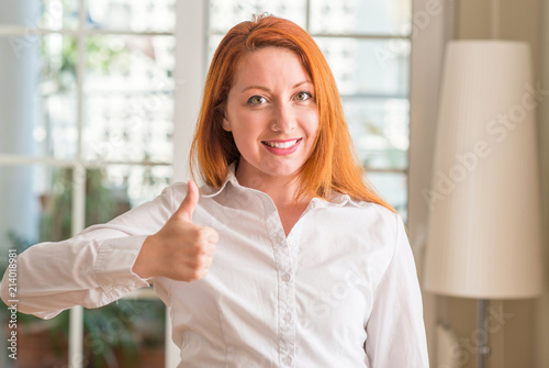 Redhead woman wearing white shirt at home happy with big smile doing ok sign, thumb up with fingers, excellent sign