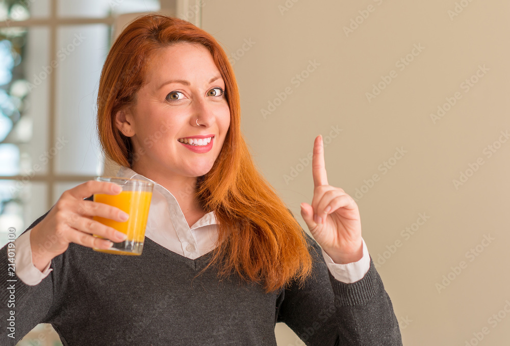 Redhead woman holding orange juice glass at home, surprised with an idea or question pointing finger with happy face, number one Redhead woman holding orange juice glass at home, surprised with an idea or question pointing finger with happy face, number one