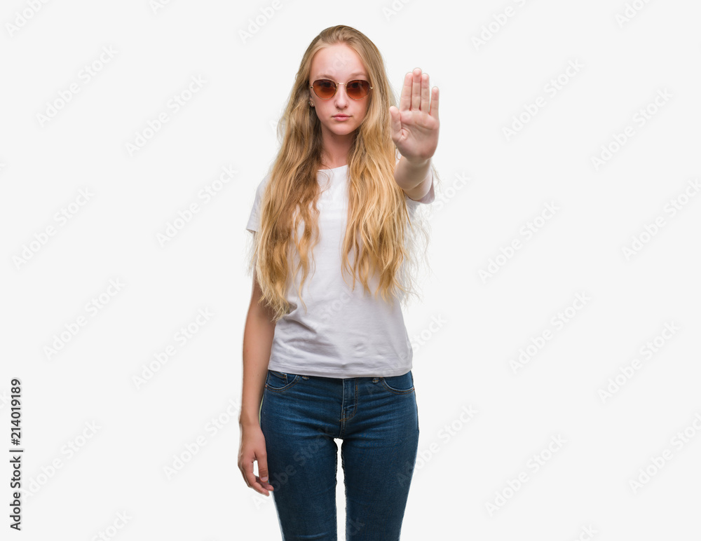 Blonde teenager woman wearing sunglasses with open hand doing stop sign with serious and confident expression, defense gesture Blonde teenager woman wearing sunglasses with open hand doing stop sign with serious and confident expression, defense gesture