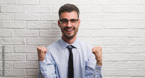 Young adult business man standing over white brick wall screaming proud and celebrating victory and success very excited, cheering emotion