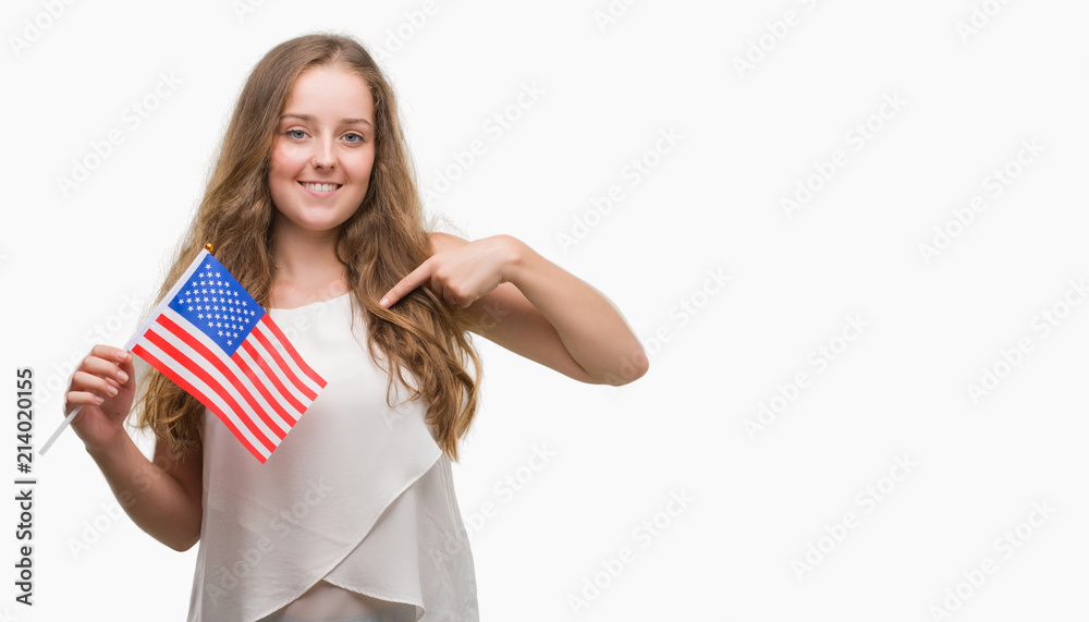 Young blonde woman holding flag of USA with surprise face pointing finger to himself Young blonde woman holding flag of USA with surprise face pointing finger to himself