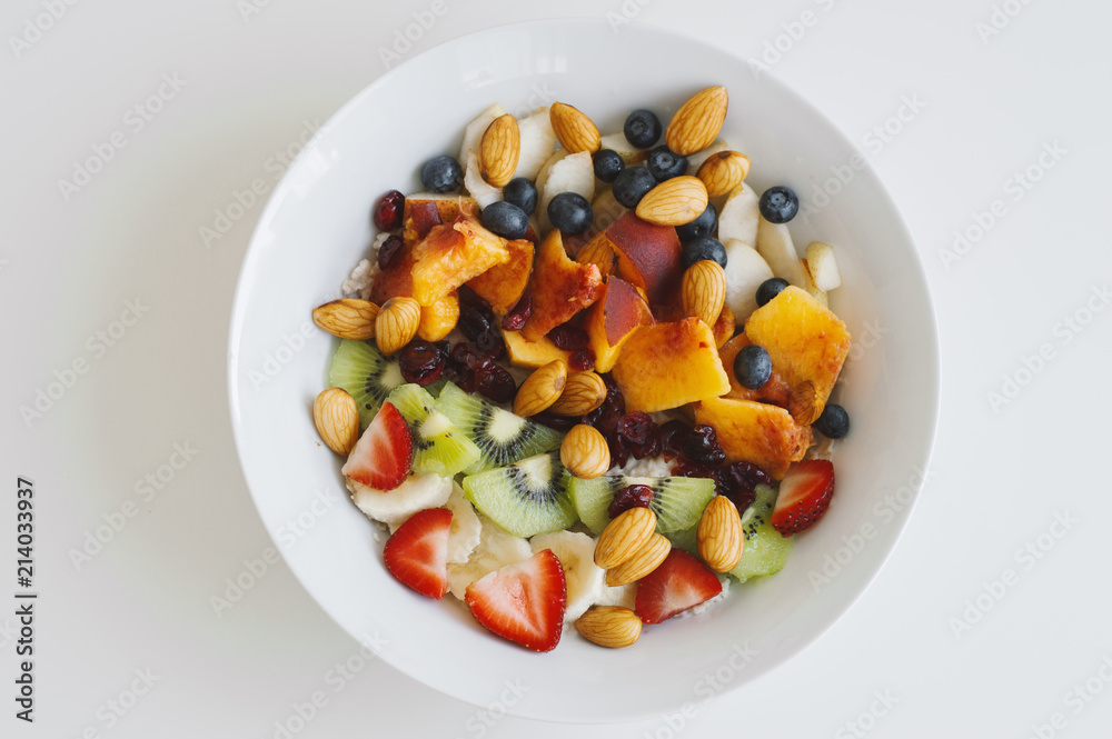 White bowl with healthy and sweet breakfast: oats, strawberry, kiwi, peach, banana, dried cranberries, blueberry and almond, top view