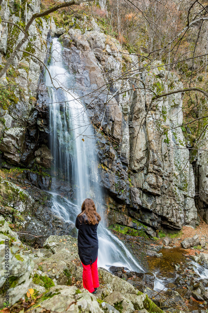Blurred long exposure photo, Boyanski waterfall, Boyana river near Sofia, Bulgaria in fall, cold autumn day and beautiful girl enjoying nature in tourist clothes, black jacket and red trousers