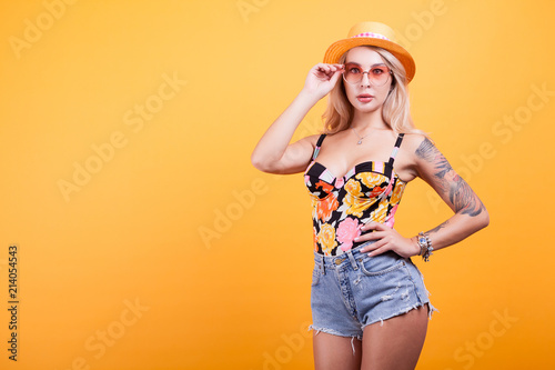 Young sexy slim tanned woman in swimsuit with sunglasses and hat over yellow background