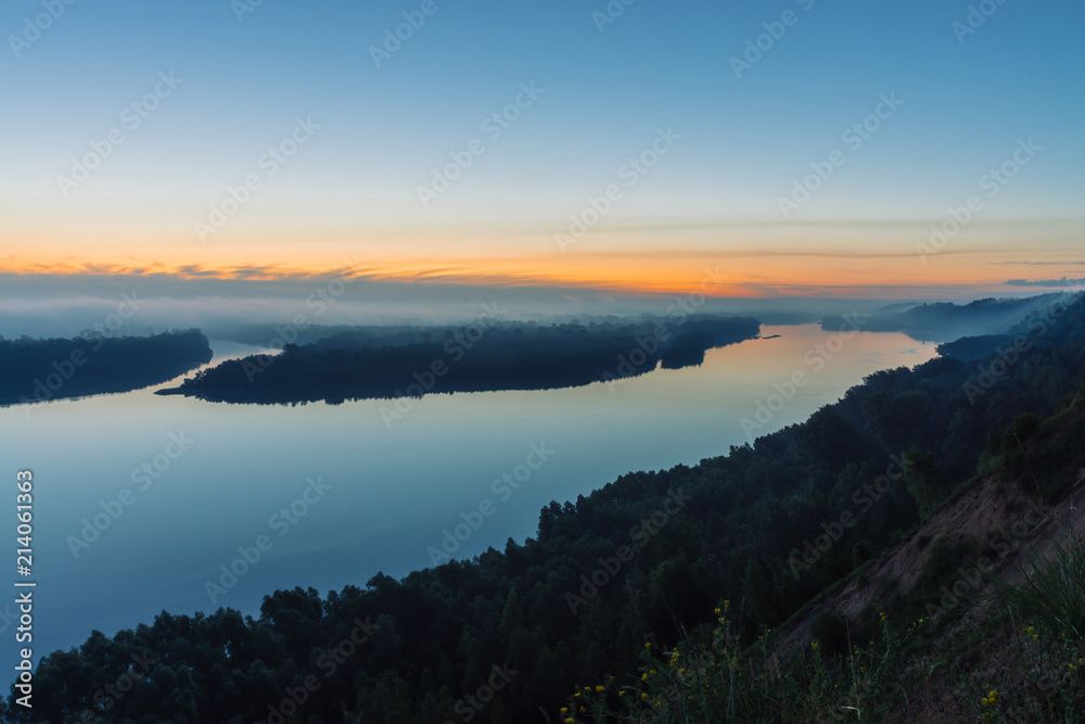 View from high shore on river. Riverbank with forest under thick fog. Dawn reflected in water. Yellow stripe in picturesque predawn sky. Colorful calm morning atmospheric landscape of majestic nature.