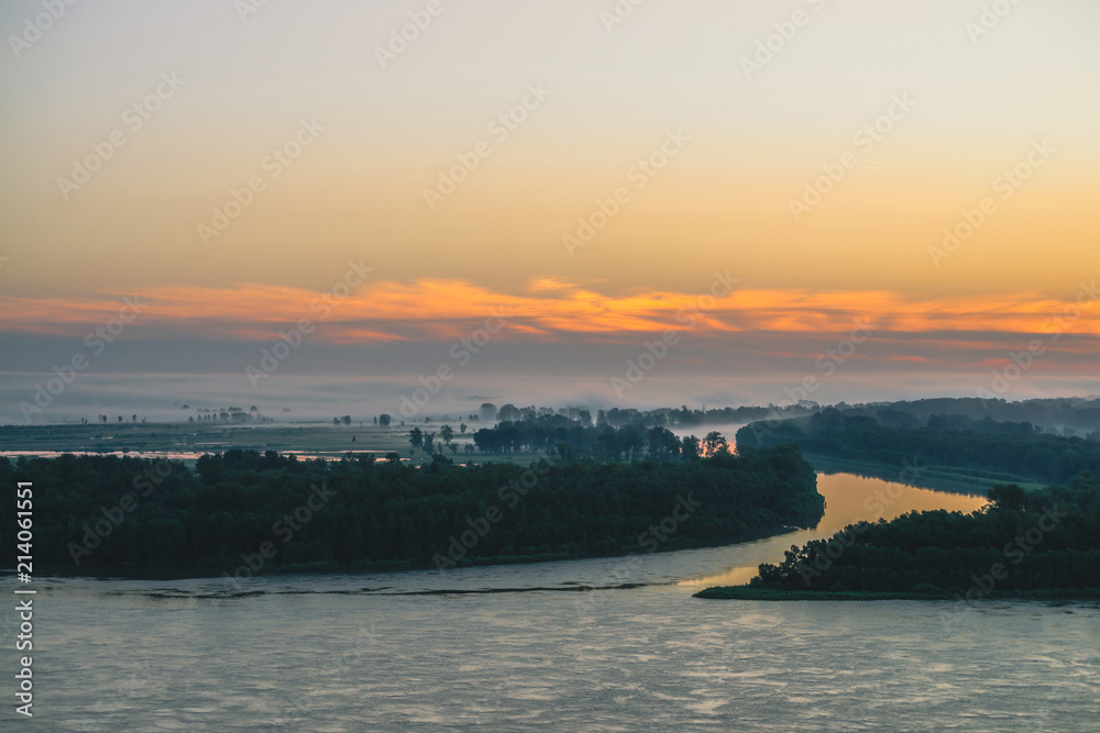 Early blue sky reflected in river water. Riverbank with forest under predawn sky. Orange stripe in picturesque sky. Fog hid trees on island. Colorful morning atmospheric landscape of majestic nature.