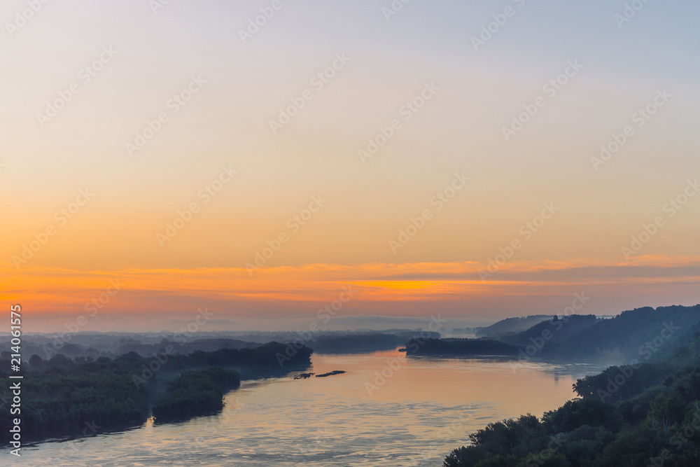 View from high shore on river. Riverbank with forest under thick fog. Gold dawn reflected in water. Orange glow in picturesque predawn sky. Mystical morning atmospheric landscape of majestic nature.