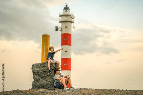 Two smiling brothers sitting near a lighthouse at the sunset with a cloudy sky in background. The youngest indicates the horizon with a finger. Two young boys with glasses sitting on the rocks