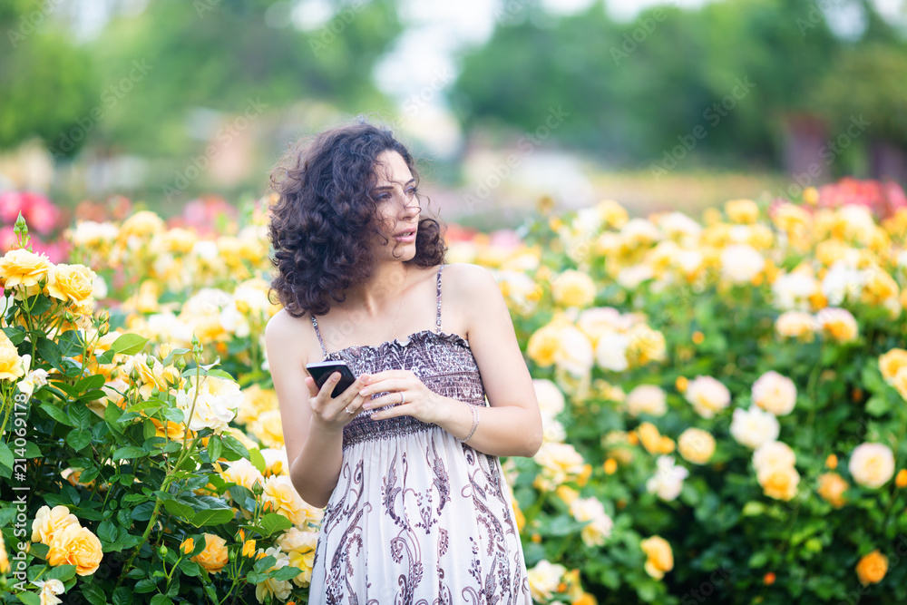 Young Caucasian woman with dark curly hair near yellow rose bush in a rose garden with black smartphone, reading emails, checking messages, looks to her left Young Caucasian woman with dark curly hair near yellow rose bush in a rose garden with black smartphone, reading emails, checking messages, looks to her left