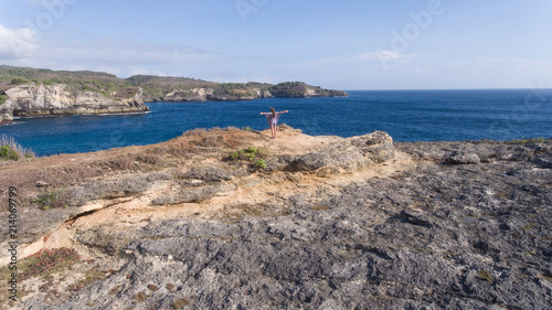 Aerial view of young girl stands on the edge of a cliff and looks at the sea Nusa Penida. Girl raising her hands up on the edge of the cliff enjoys the view of the ocean. Travel concept.