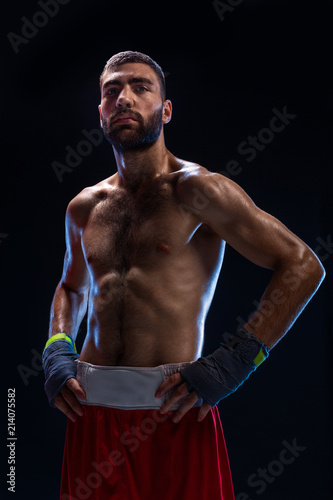 Handsome athletic guy in a red shorts on a black background. The boxer is fetching his breath after practicing hooks and blows. © nazarovsergey