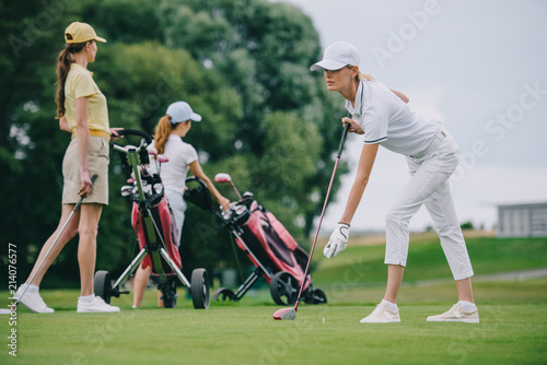 Selective focus of focused woman in cap playing golf with friends near by at golf course