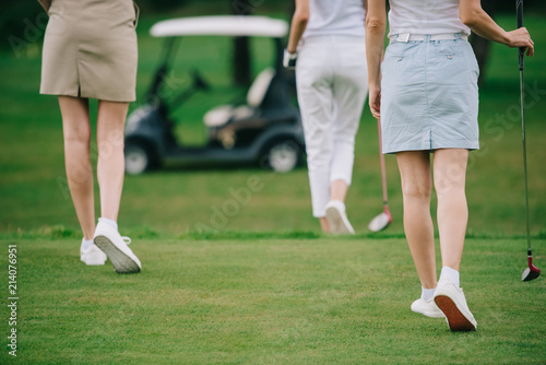 cropped shot of women with golf clubs walking on green lawn at golf course