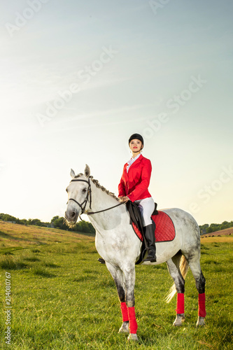 Obraz na plátně Young woman rider, wearing red redingote and white breeches, with her horse in evening sunset light