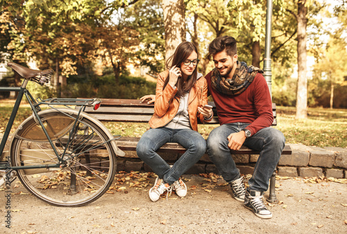 Young couple sitting outdoors at the park on beautiful autumn day.They sitting on bench and using smart phone.