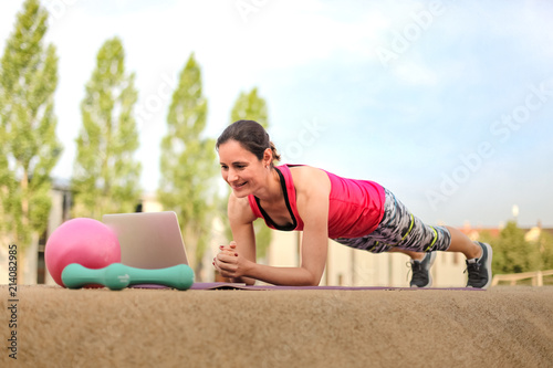 sporty and strong woman making planks infront of a laptop while watching online Fitness program on a mat in the park photo