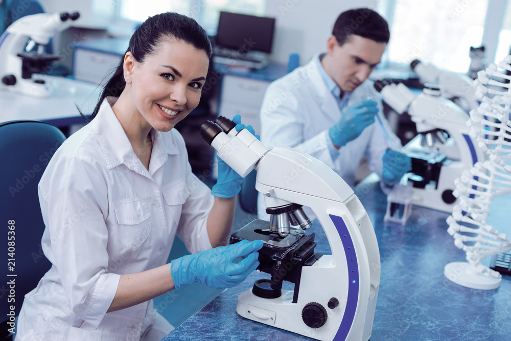 Modern genomics. Delighted positive female geneticist smiling and looking at you while working in the lab with the microscope Modern genomics. Delighted positive female geneticist smiling and looking at you while working in the lab with the microscope
