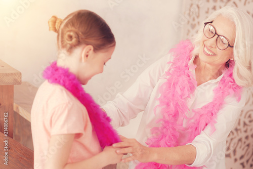 You are so cute. Selective focus on a beautiful retired lady smiling broadly while looking at her adorable granddaughter wearing a feather boa while both having fun at home.