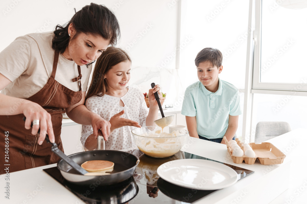 Adult brunette woman and happy children 8-10 cooking together, and frying pancakes on modern stove in kitchen at home