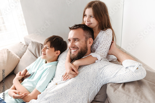 Happy father 30s with son and daughter hugging together on sofa at home, and looking aside