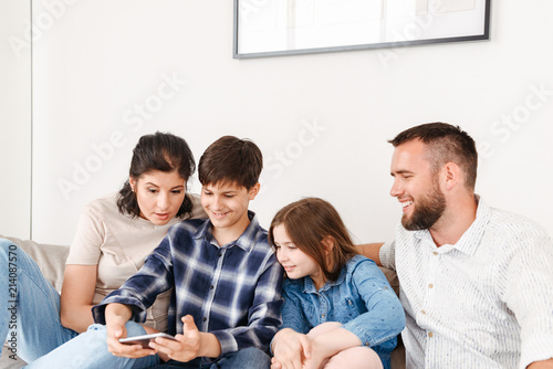 Image of caucasian family with two children sitting on sofa together at home, and using smartphone